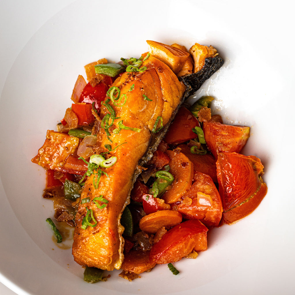 A close-up shot of a cooked salmon fillet served over a bed of roasted tomatoes, onions, and green peppers in a white bowl.