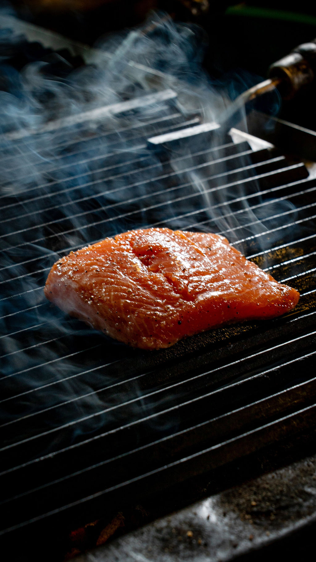 A raw salmon fillet cooking on a dark grill grate with visible smoke rising around it.